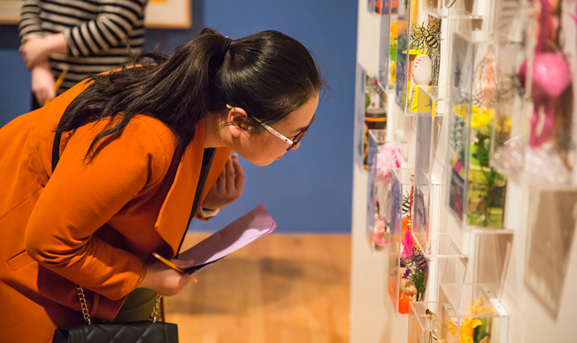 student with glasses and floral sweater examining artwork mounted to wall