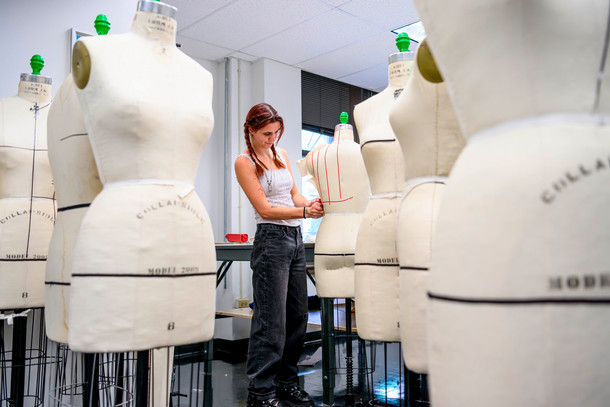 Standing among a sea of dress forms of different sizes, a student maps out a pattern in red. 