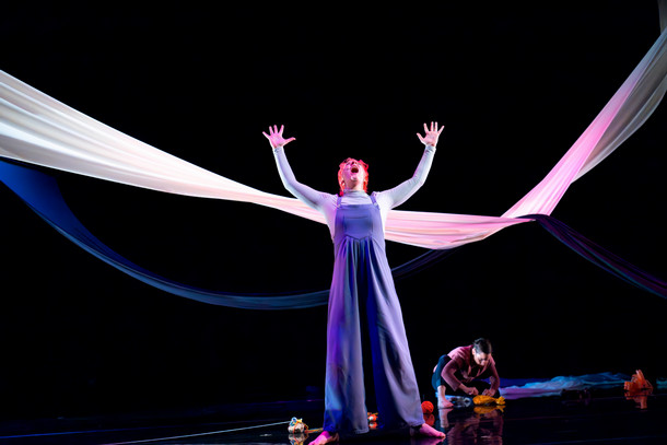 A performer on stage raises their arms and shouts toward the sky on stage. A large ribbon stretches across the stage above them.