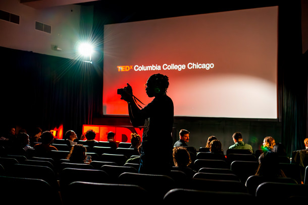 A student with a camera is silhouetted in a dark room. Behind them is a red lit up presentation screen with "TEDX Columbia College Chicago" displayed. 