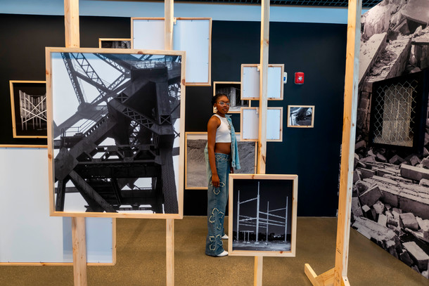 A wooden housing frame holds up a variety of black and white pieces of artwork. A student stands within the framing looking at the art