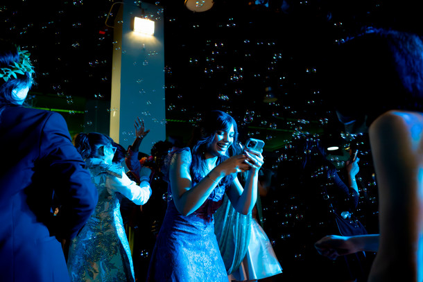 In a dark crowded room, a woman bathed in green light and surrounded by bubbles uses her cell phone and smiles.