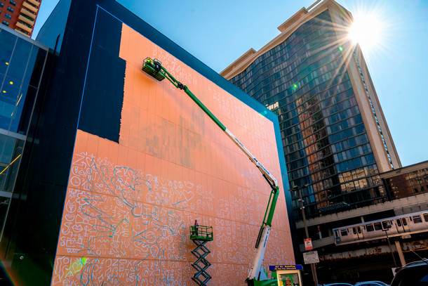 A lift carries artists high in the air as the work on an orange and white base to a mural on the side of the student center. The sun shines out from the buildings in the background.