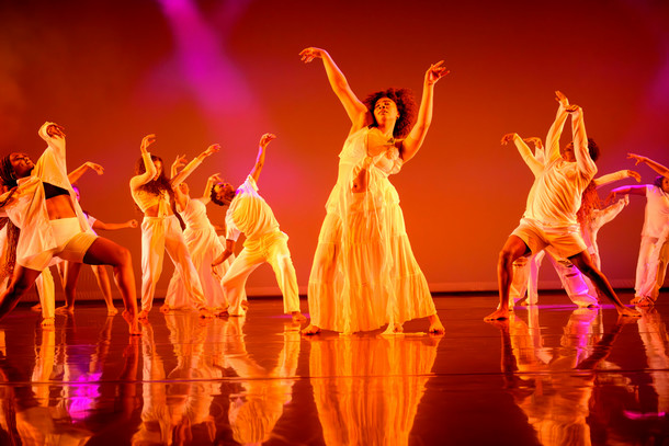 A group of students dance together on stage bathed in orange light. 