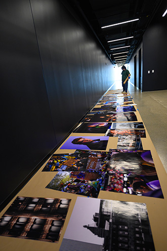 A student stands in the middle of a long hallway looking down at photo prints laying organized on the floor before they are put on the wall. 