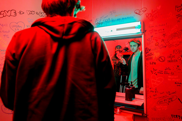 In a movie set of a bathroom bathed in red light, film students work on a horror film, graffiti covers the walls, and two people look into a mirror.