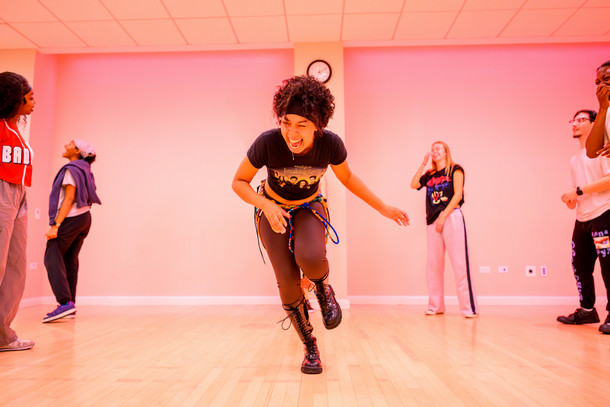 A group of students dance in a a group in a pink lit room. The student dancing in the middle smiles widely and the others look on in joy.