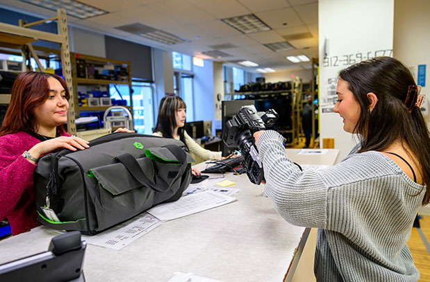 student at desk getting camera with two other students on other side of desk