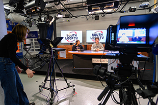 students standing behind cameras filming students sitting behind news desk