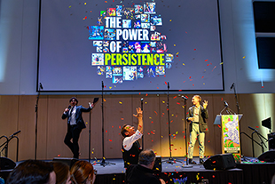 two people on stage with screen in background with text power of persistence and person in foreground throwing confetti